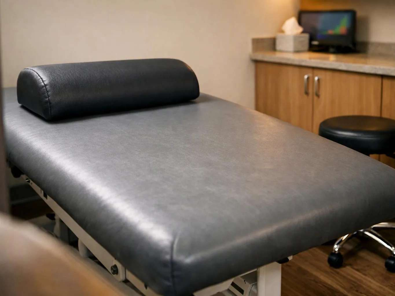 An empty exam table sits in a clinic room beside a stool, cabinets, and a tissue box