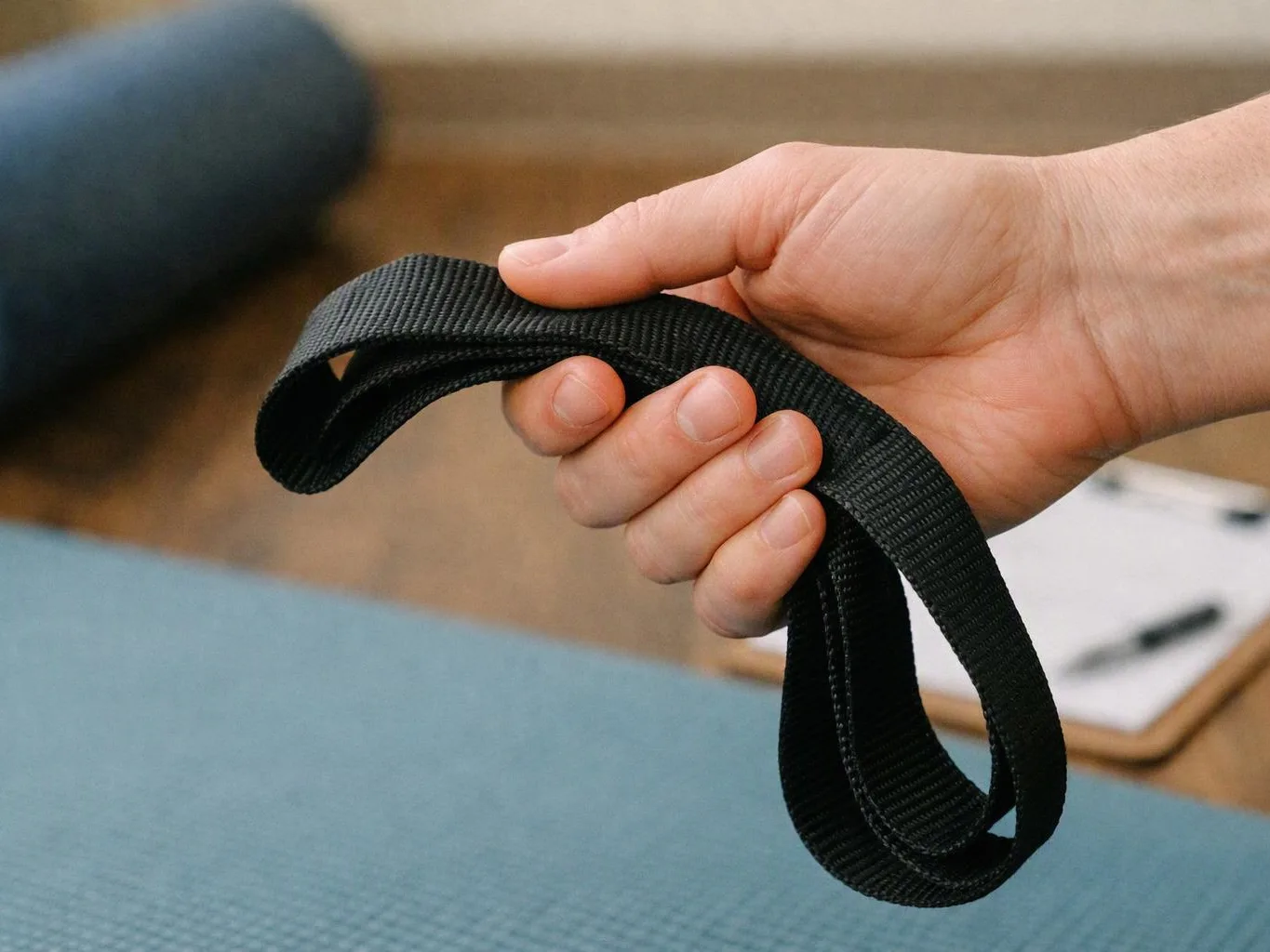 A hand holds a black woven strap over a blue exercise mat indoors