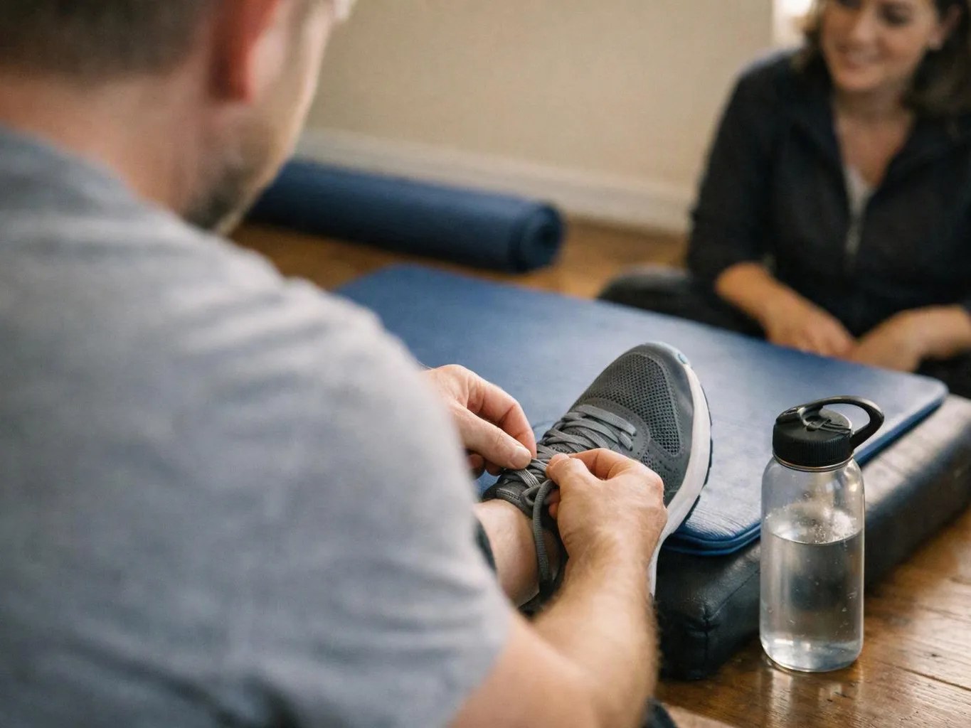 A man ties his gray sneaker while sitting on a padded mat, with a water bottle and a smiling woman nearby