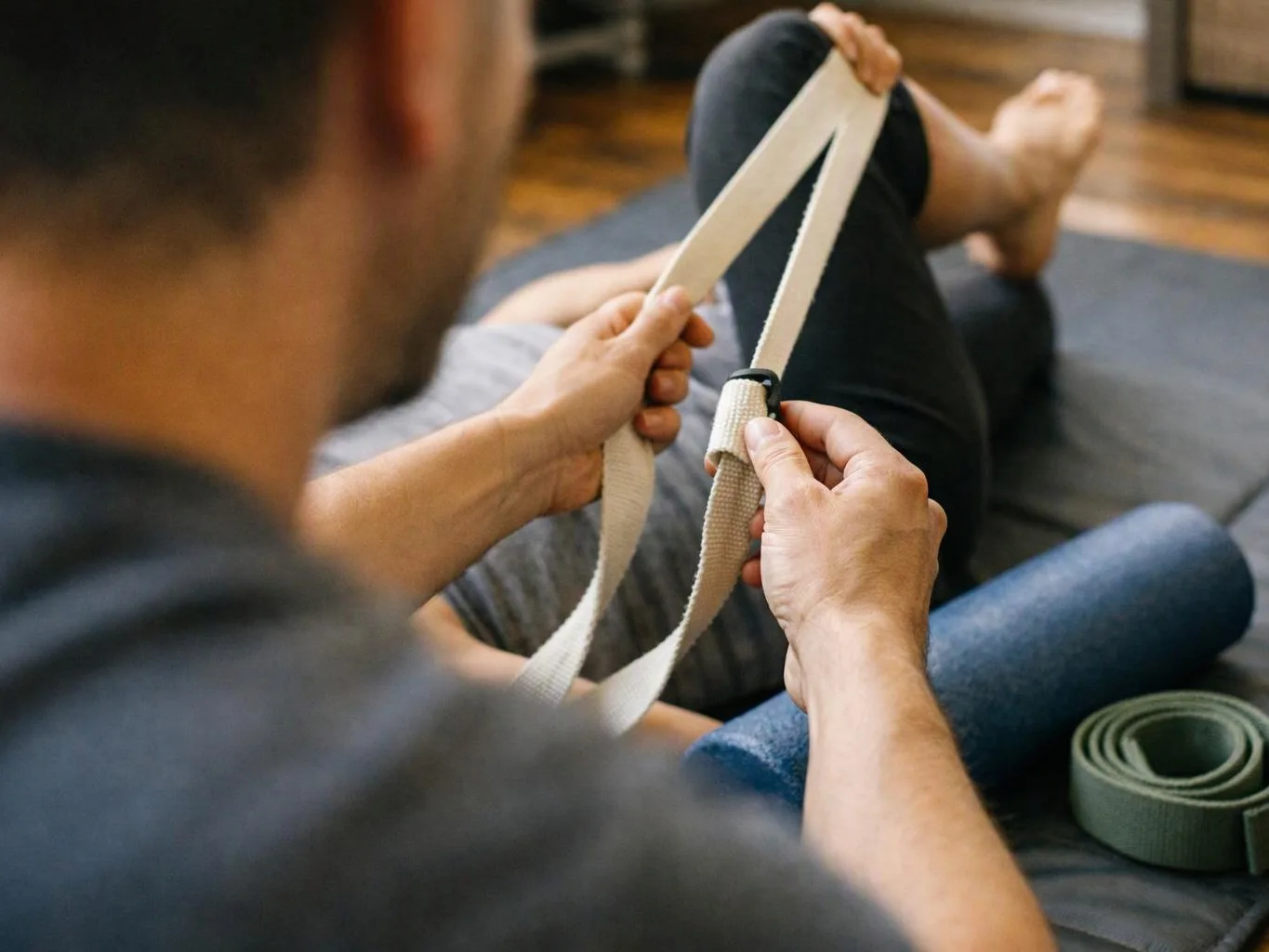 A person on a mat holds a yoga strap while stretching their leg indoors