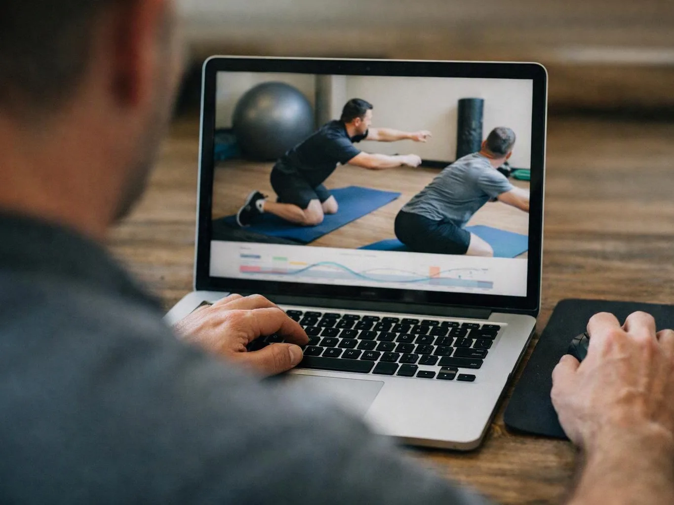 A person uses a laptop while watching a fitness video of two men exercising on mats